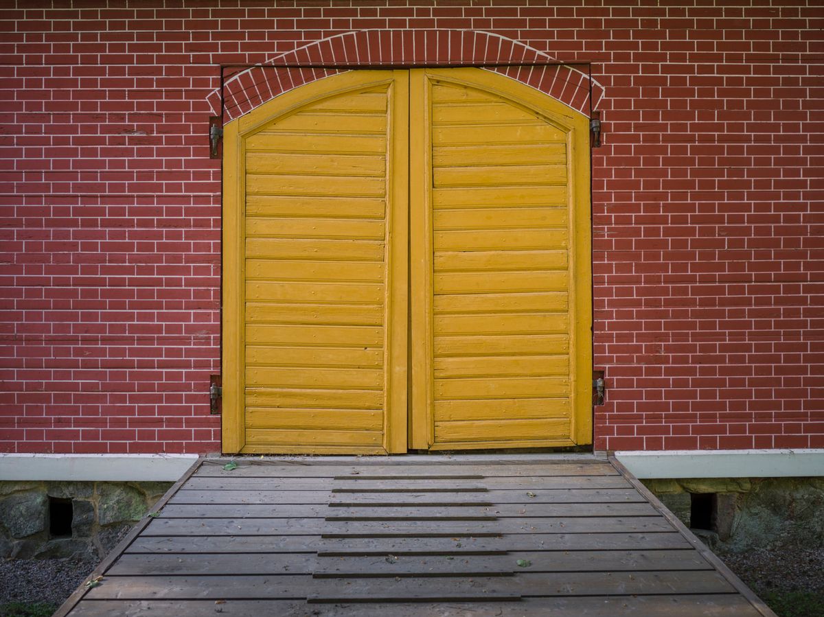 Puertas dobles amarillas en una fachada de ladrillo rojo, al final de una pasarela de madera.