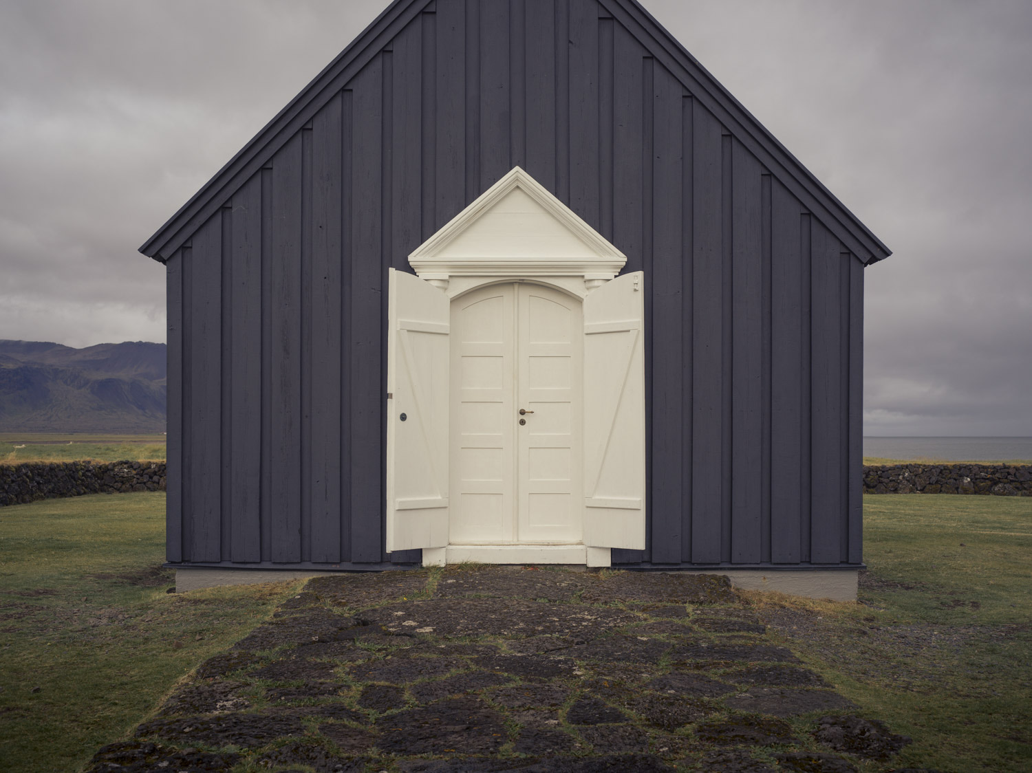 Fachada oscura a dos aguas con una puerta blanca y frontón triangular.