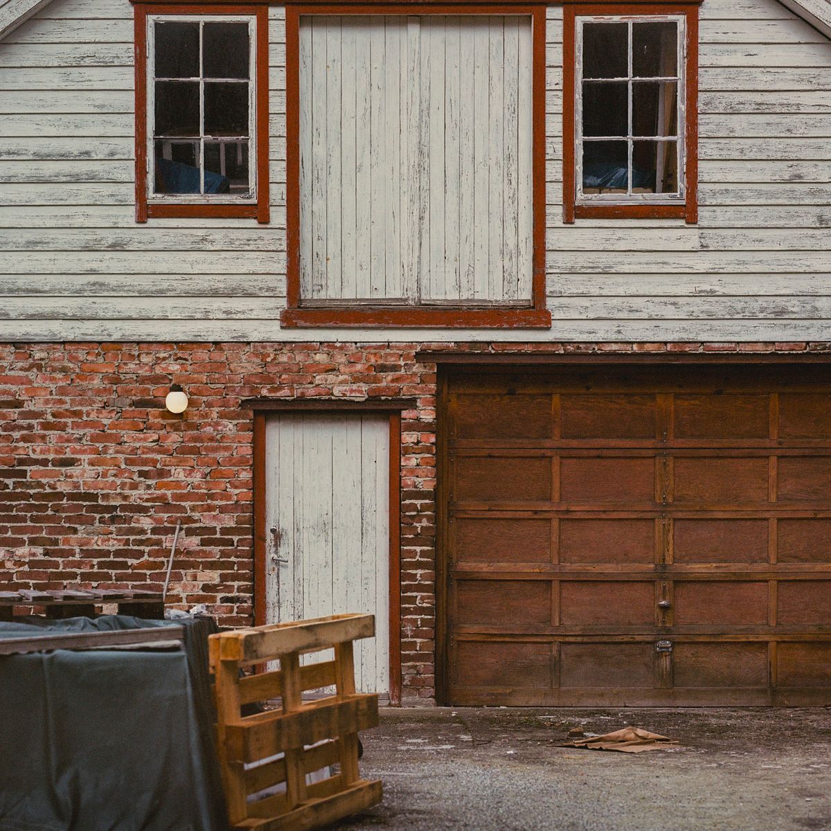 Fachada de madera y ladrillo con puertas tapiadas, portón marrón y un palé.