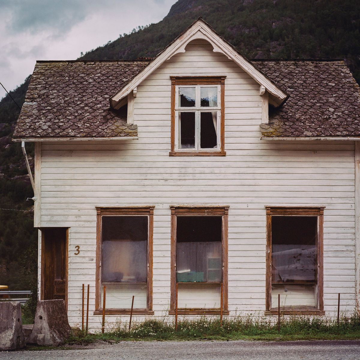 Casa de madera blanca con tejado a dos aguas y tres ventanas inferiores, ante una montaña.