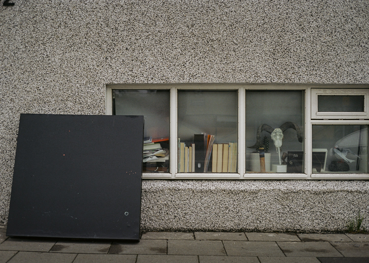 Panel negro apoyado contra una fachada, junto a una ventana baja con libros y objetos.