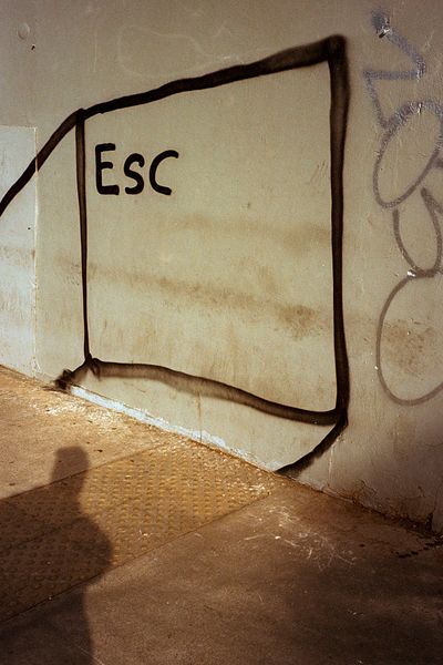 Dibujo de una tecla «Esc» en una pared clara, con una sombra humana sobre la acera