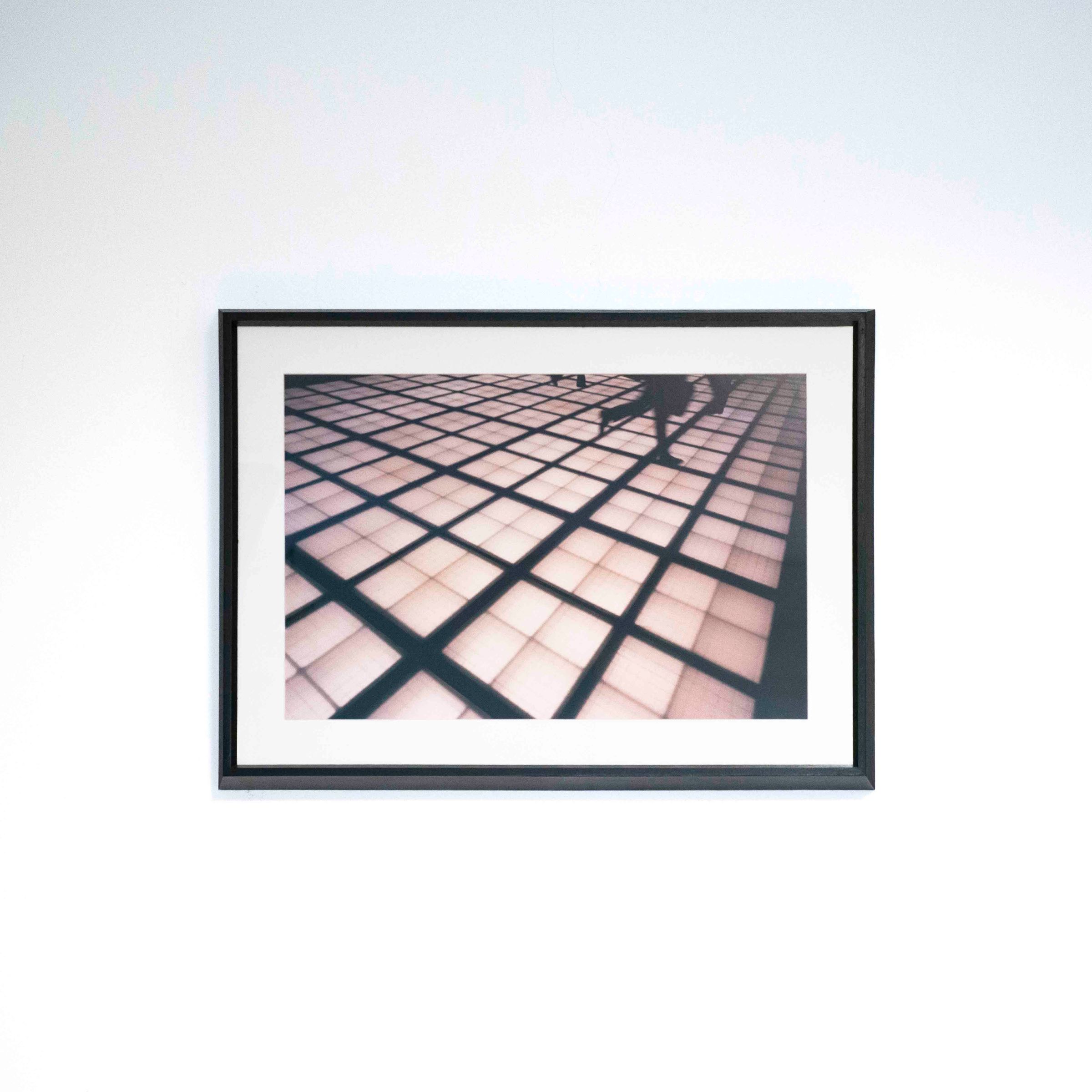 Fotografía horizontal de una cuadrícula de sombras y piernas caminando, con marco negro y paspartú blanco sobre una pared blanca.