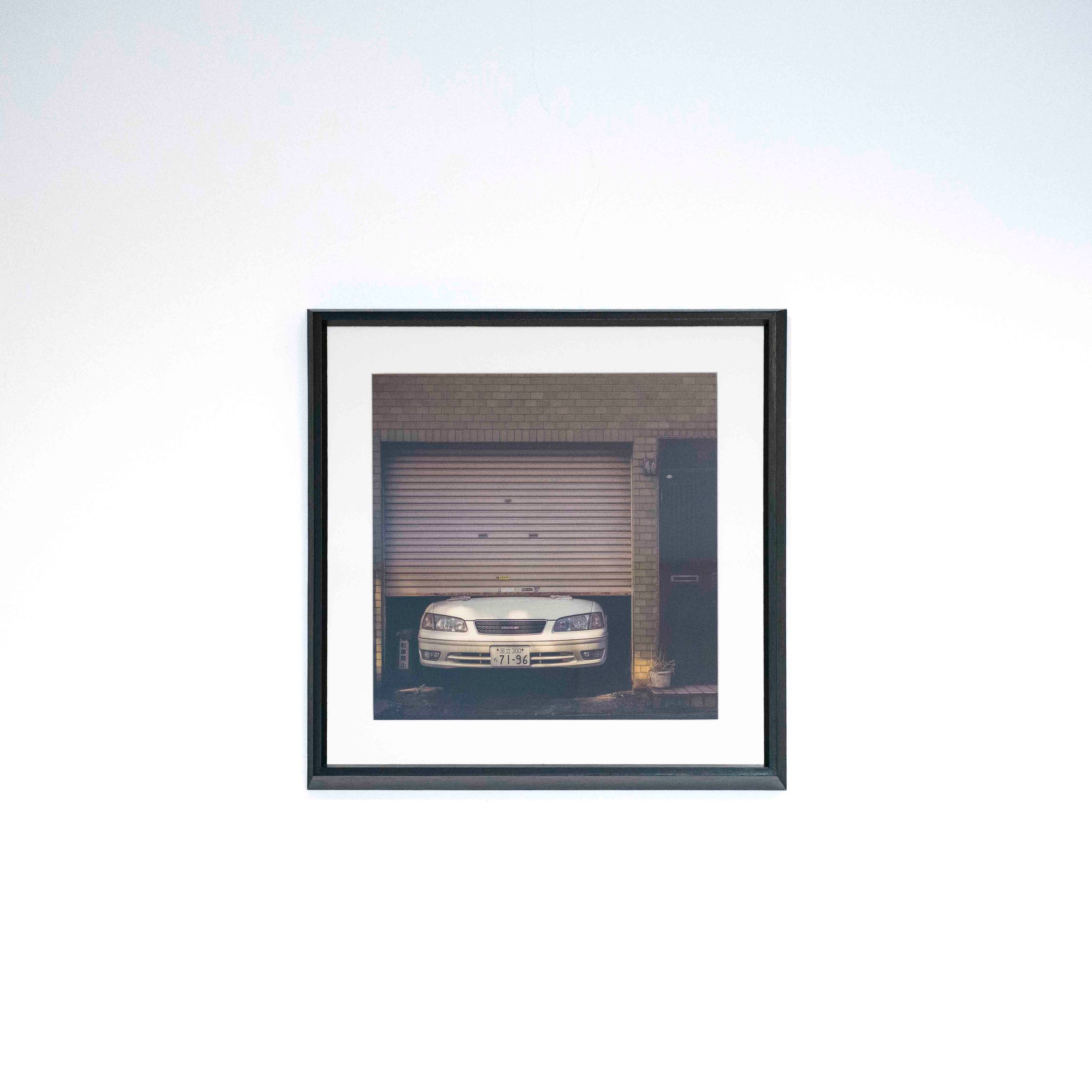 Fotografía de un auto bajo una persiana metálica, con marco negro y paspartú blanco sobre una pared blanca.