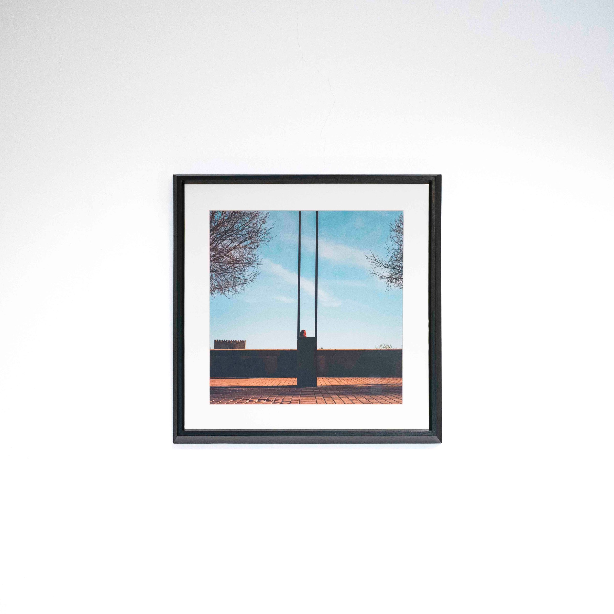 Fotografía de una figura entre postes altos, árboles y cielo azul, con marco negro y paspartú blanco sobre una pared blanca.