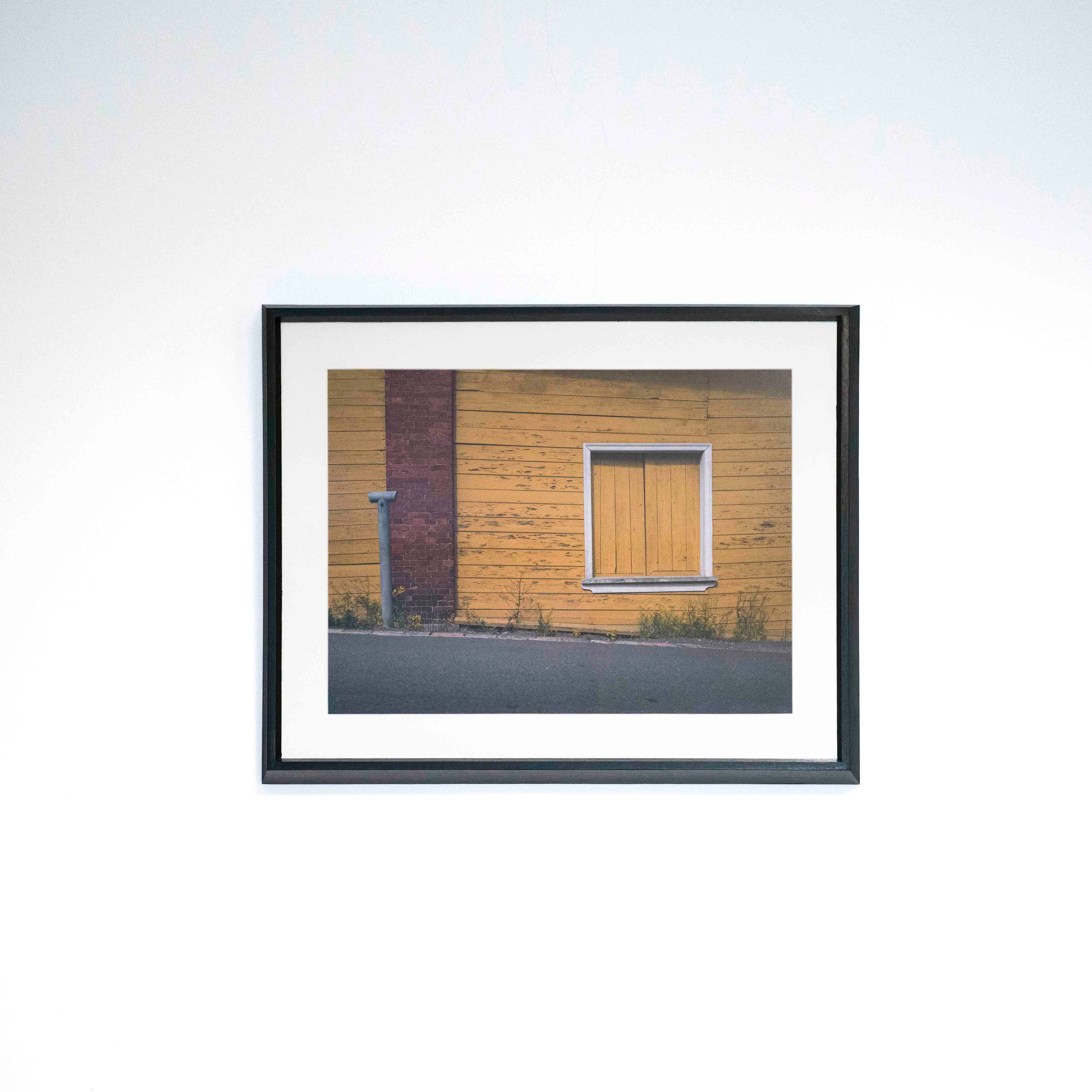 Fotografía de una ventana tapiada en una pared amarilla, con marco negro y paspartú blanco sobre una pared blanca.