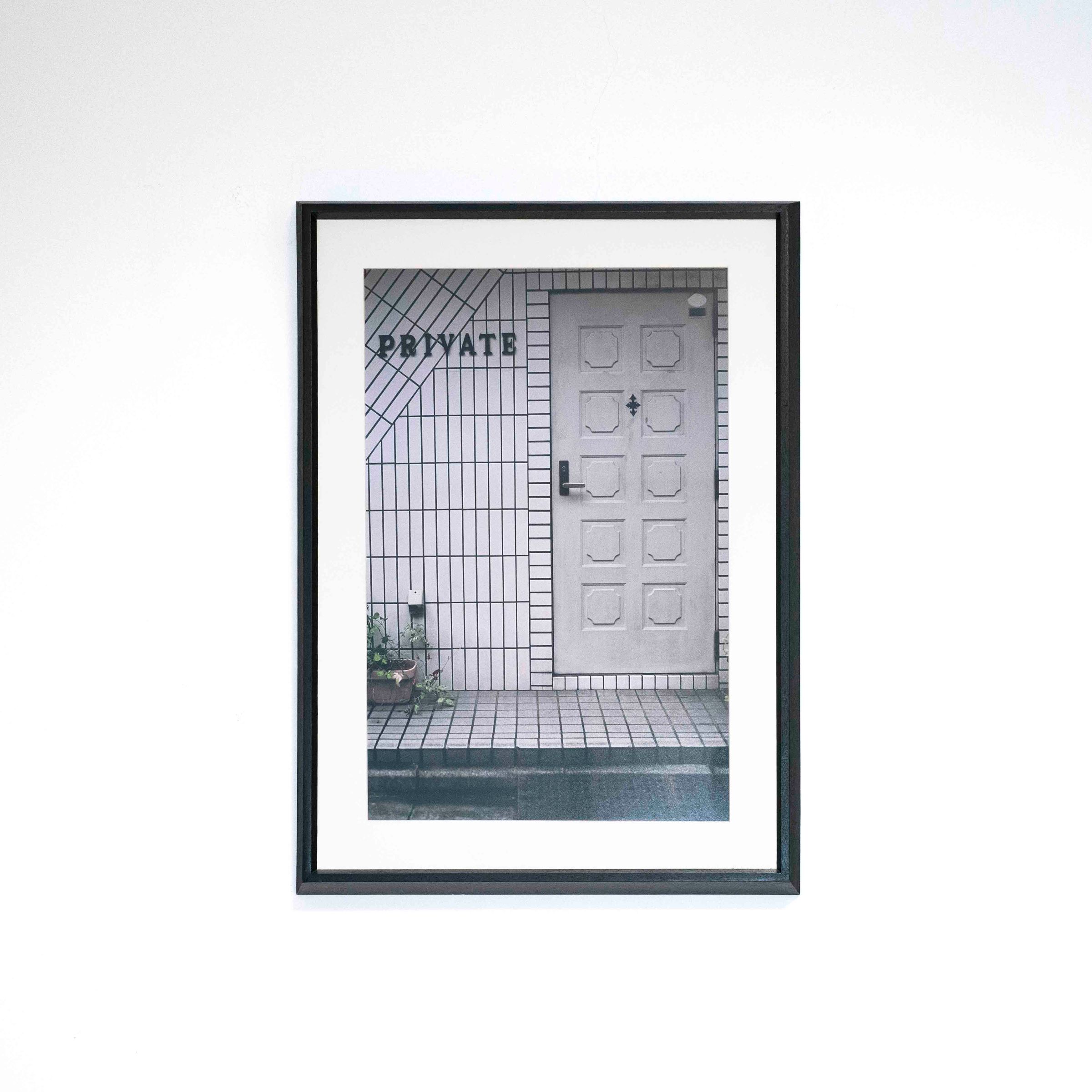 Fotografía vertical de una puerta junto a la palabra «PRIVATE», con marco negro y paspartú blanco sobre una pared blanca.