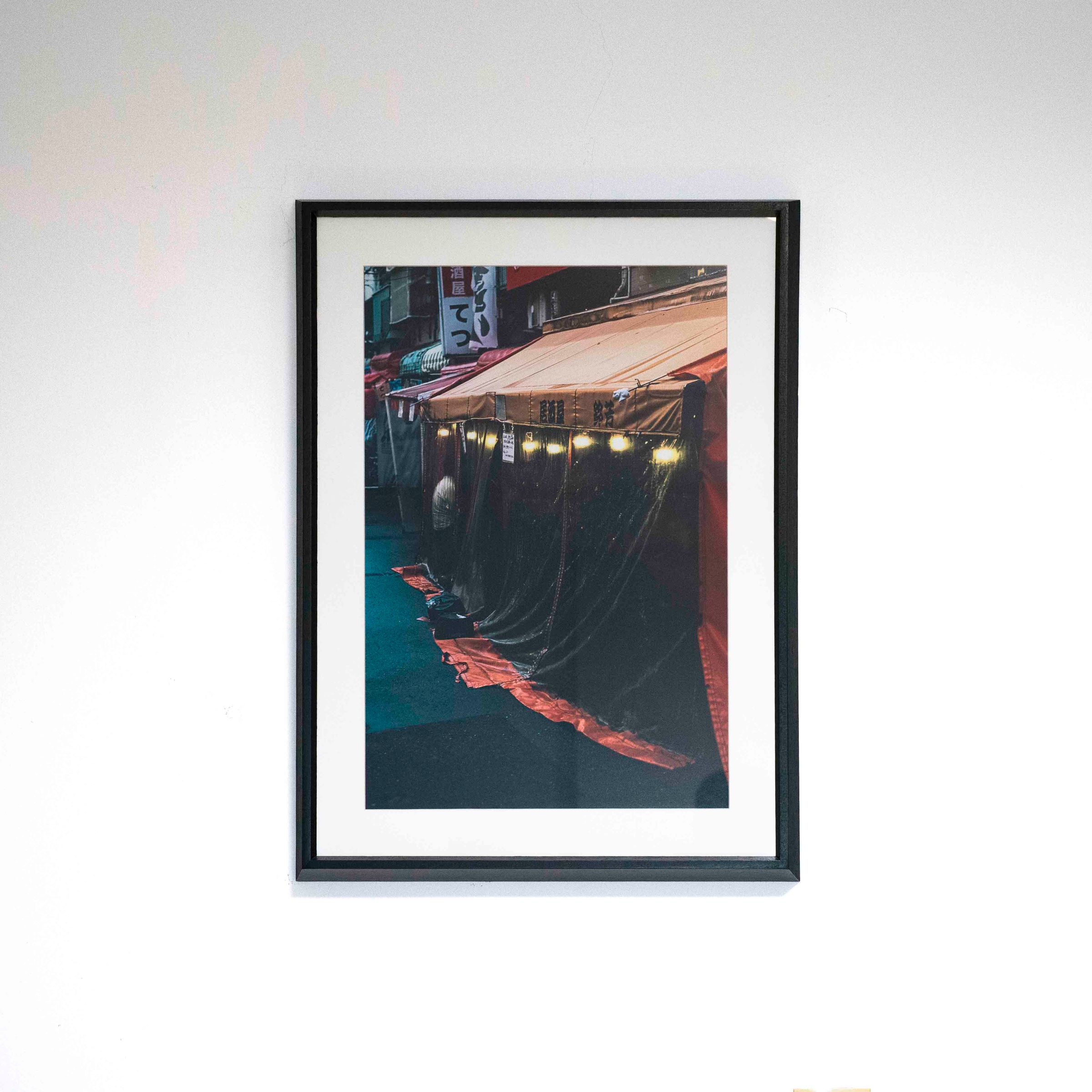 Fotografía vertical de un local con toldo y cortinas oscuras, con marco negro y paspartú blanco sobre una pared blanca.