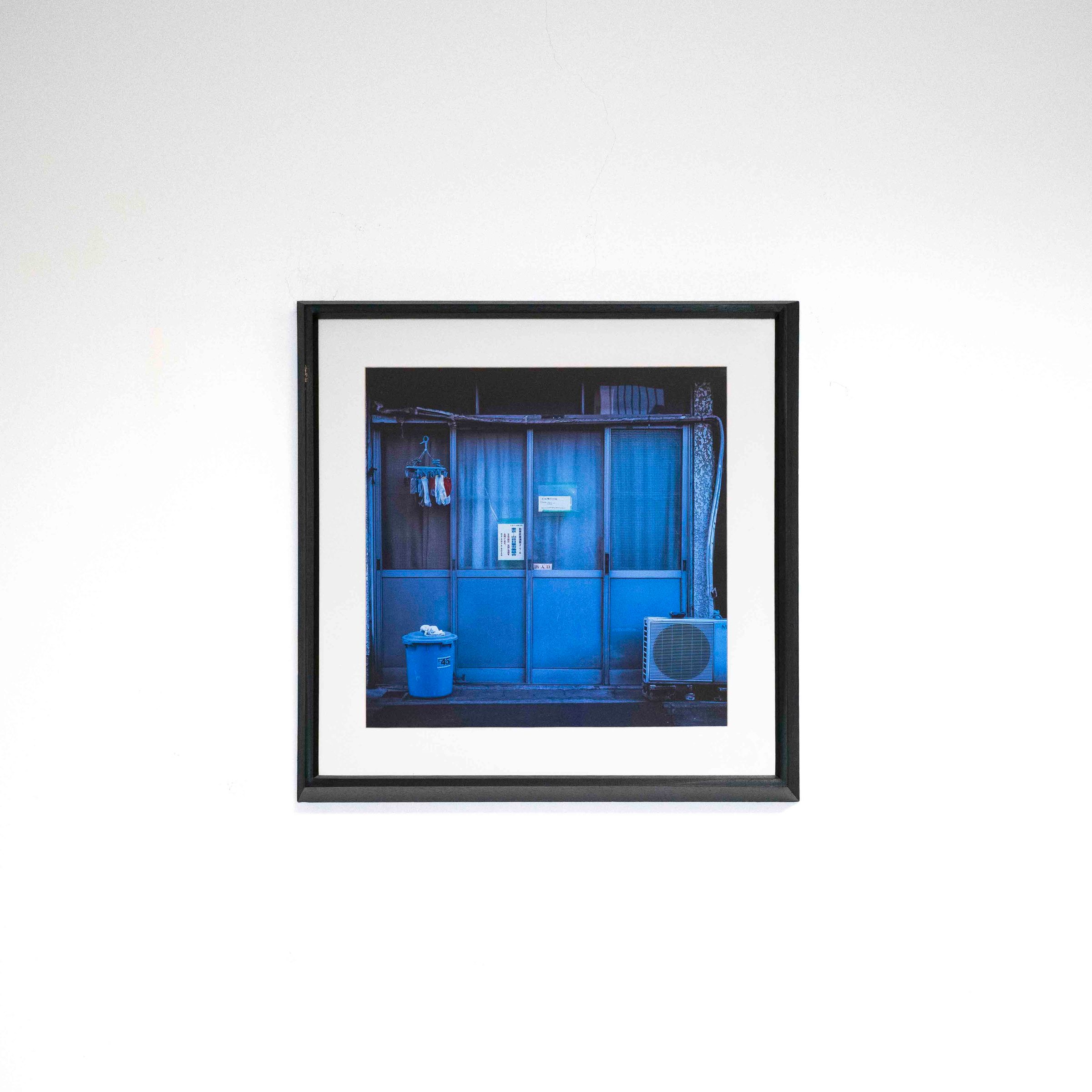 Fotografía de una entrada azul con cortinas, un balde y un equipo de aire acondicionado, con marco negro y paspartú blanco sobre una pared blanca.