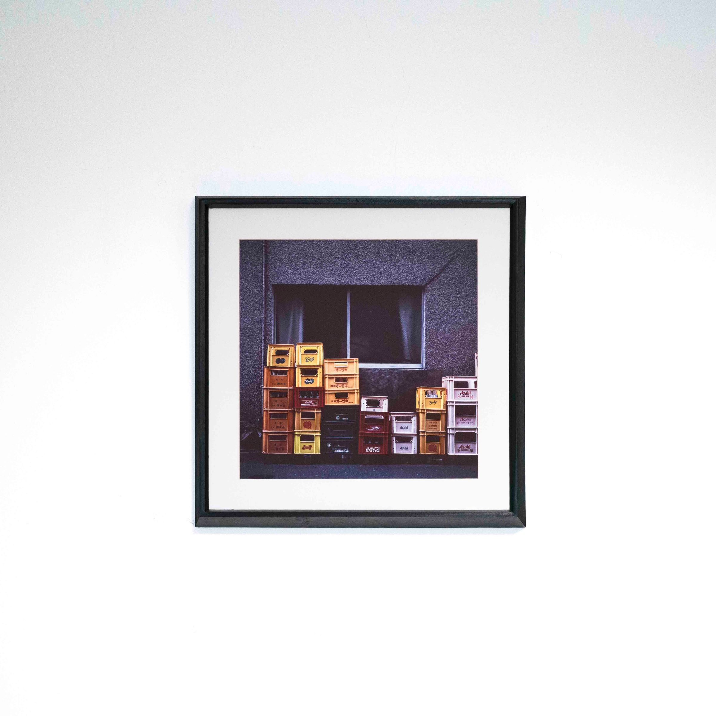 Fotografía de cajones de colores apilados bajo una ventana, con marco negro y paspartú blanco sobre una pared blanca.