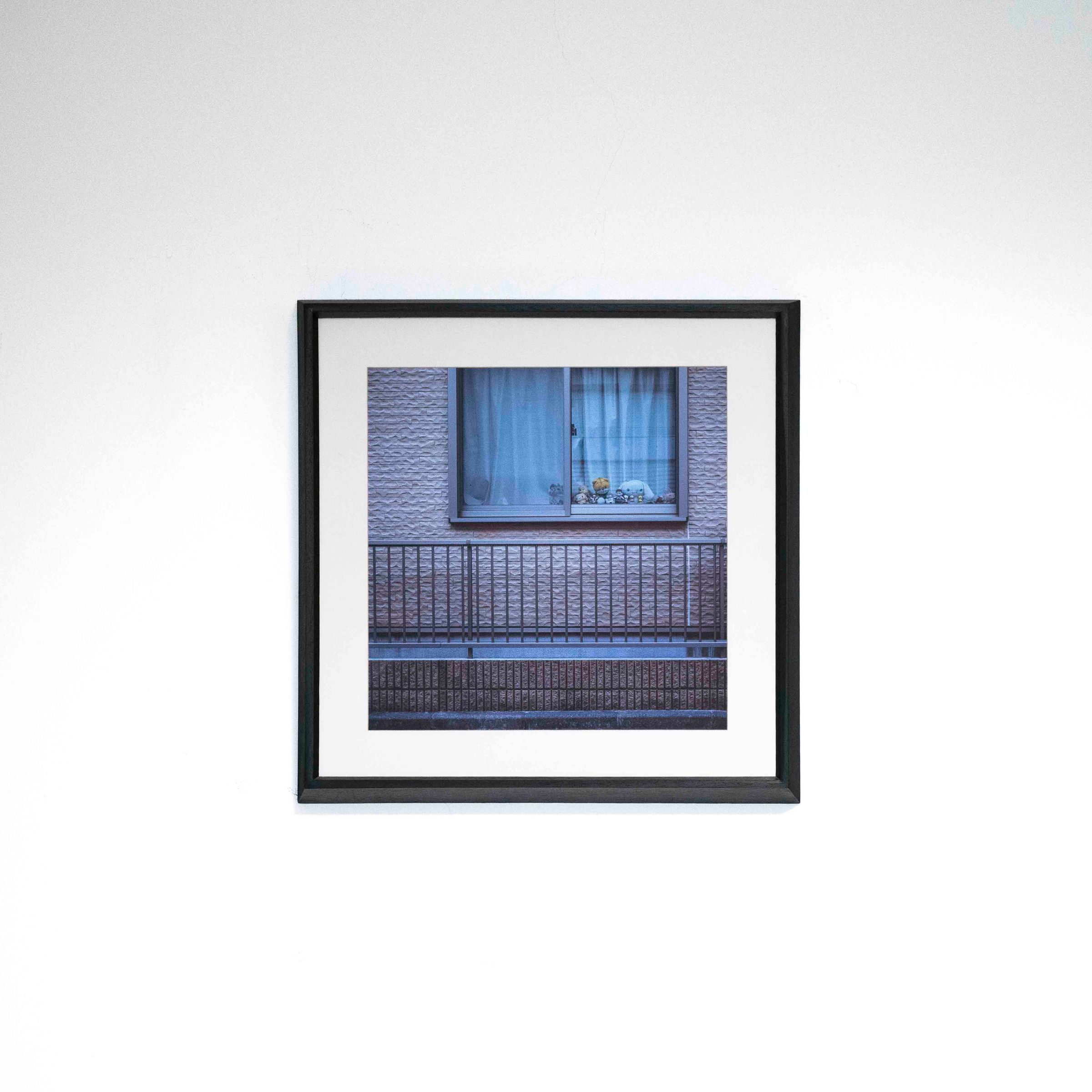 Fotografía de una ventana con cortinas azules detrás de una baranda, con marco negro y paspartú blanco sobre una pared blanca.
